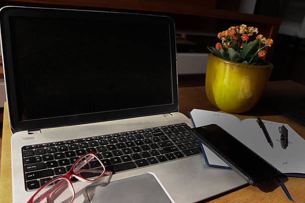 Cozy home office desk setup with a laptop, yellow coffee mug, pink-framed reading glasses, a stack of books, and a kalanchoe flower in a mustard yellow vase. Bright and creative remote workspace with natural light and warm tones.