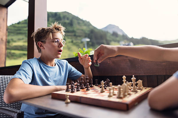 Two people are engaged in a thoughtful chess game on a wooden deck or patio with a breathtaking view of rolling green hills and distant mountains. The boy is realizing that he made a mistake and the opponent is going to win.Shot with Canon R5