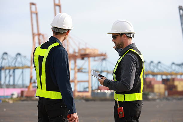 istock Two men with green safety vests and PPE are working at the port, using the logistics export-import transportation concept.  Two males are working in a logistics shipping yard. Marine and carrier insurance concept. Cargo container yard. 2213893276