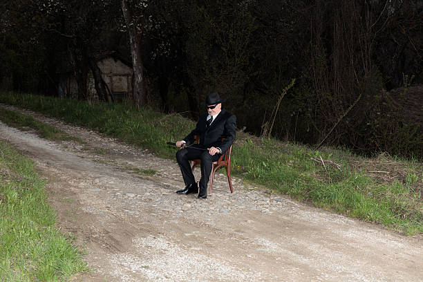 Man in formal suit, tie, sunglasses, bowler hat holding katana, on country road