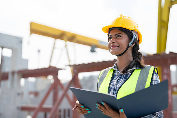 istock Asian woman engineer holding document smiling at construction site. Confident female Indian wearing protective helmet and vest working in factory making precast concrete wall for real estate housing. 2213913084
