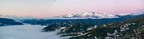 A stunning panoramic vista captures the majestic, snow-capped peaks of the Caucasus Mountains near Stepantsminda, Georgia, bathed in the soft, pastel hues of dawn. A sea of fog or low clouds fills the valleys below, creating a dramatic and ethereal landscape. The first light of day illuminates the snow-covered summits, contrasting with the darker, forested slopes and the misty depths, evoking a sense of awe and the vastness of the mountain range.