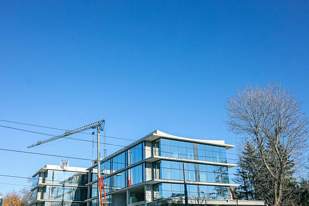 Modern glass office building under construction with a tower crane on a clear blue sky background, surrounded by bare trees and power lines in an urban setting.