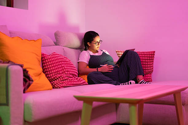 istock Pregnant woman relaxing on sofa using digital tablet in pink lit room 2213923866