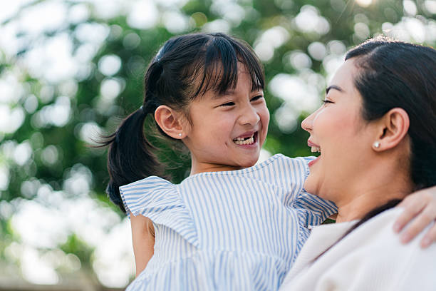 A joyful moment is shared between an Asian mother and her young daughter as they laugh together in an outdoor setting. The child, dressed in a light blue striped dress with flutter sleeves, is lifted in her mother’s arms and beams with happiness. The sunlight shines through the trees behind them, enhancing the warm, candid atmosphere. The mother, smiling and engaged, wears a white top and gently holds her daughter close. This heartfelt image captures the essence of love, family bonding, and the beauty of authentic connections in everyday life.
