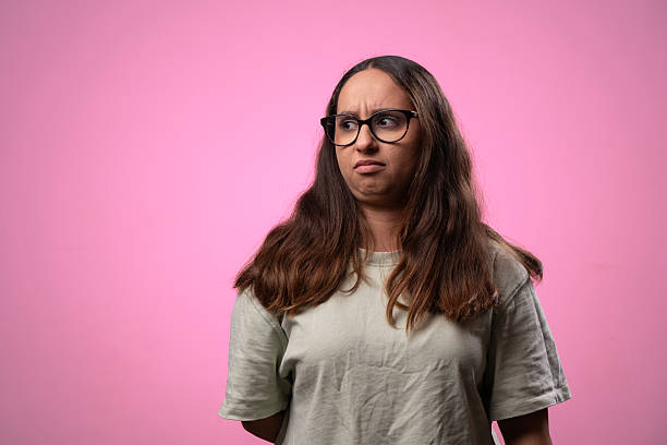 Studio shot of a young woman with long brown hair and eyeglasses grimacing and looking away, conveying feelings of disgust and skepticism against a vibrant pink backdrop