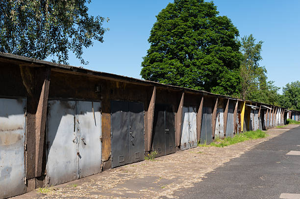 A long line of weathered metal garage doors under a clear blue sky, flanked by tall trees. The scene captures a typical Eastern European suburban atmosphere with signs of age and utilitarian design.