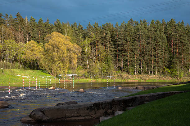 A scenic forest river with whitewater slalom poles, rocky stream, and lush greenery under a dramatic cloudy sky, ideal for kayaking or canoe training