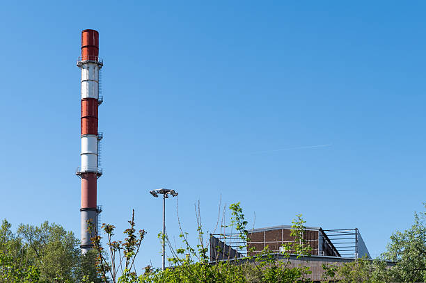 A tall red and white striped industrial smokestack rises above a partially obscured factory building surrounded by greenery. The bright blue sky and clear weather provide a stark contrast to the utilitarian structure.