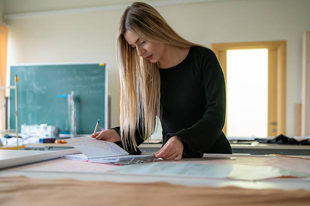 istock Student Designer focused on creating clothing designs while writing notes in a well-lit workshop filled with fabric and tools 2213950578