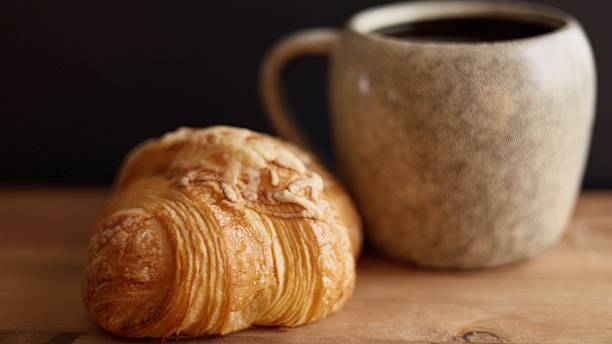 istock Croissant resting beside ceramic mug filled with black coffee on rustic wooden surface promotes artisanal breakfast. Warm, flaky pastry and rich coffee create cozy, inviting setup. 2213956650