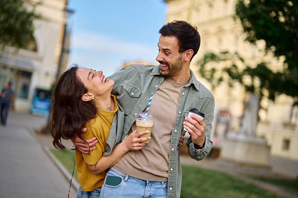 Happy young couple laughing and embracing while strolling through the city streets, enjoying their takeaway coffee and sharing moments of joy and affection together