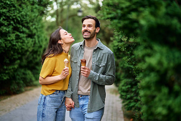 Young couple enjoying ice cream cones while walking hand in hand through a vibrant park, laughing and sharing joyful moments on a beautiful summer day filled with sunshine