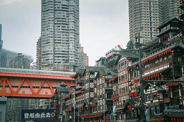 China-April 2025, Traditional stilted buildings adorned with red lanterns sit beneath a vibrant red bridge, juxtaposed against towering modern skyscrapers in Chongqing’s historic Hongyadong district.