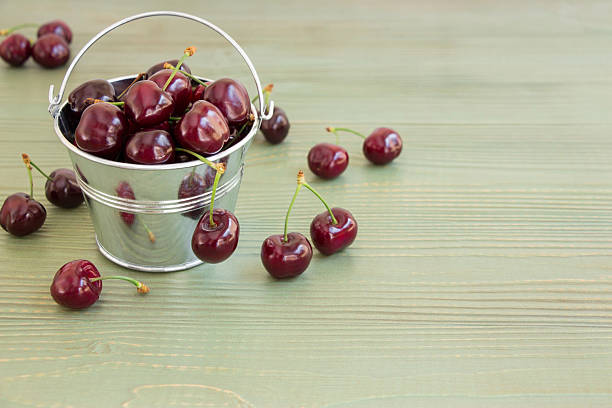 In the frame on the left a small bucket with cherries, around scatterd cherries, on the right empty space on a light green background. Bucket of cherries. Horizontal. Daylight.