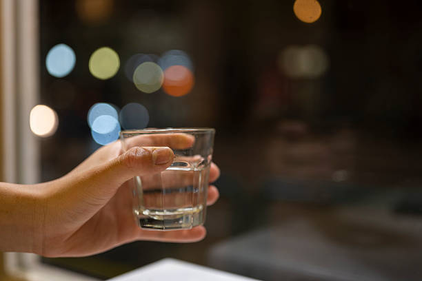 A close-up shot captures a hand gripping a glass filled with clear liquid, likely water, placed on a smooth surface. The background is softly blurred with warm bokeh lights, creating an intimate and serene atmosphere. This image could be used for themes related to refreshment, hydration, or relaxation.