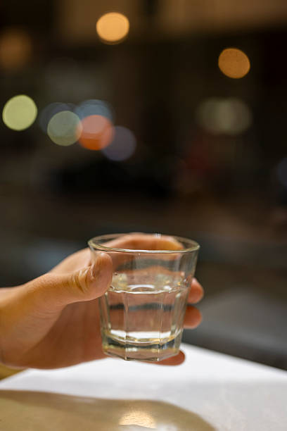 A close-up shot captures a hand gripping a glass filled with clear liquid, likely water, placed on a smooth surface. The background is softly blurred with warm bokeh lights, creating an intimate and serene atmosphere. This image could be used for themes related to refreshment, hydration, or relaxation.