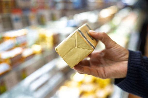 Close-up of a hand holding a wrapped block of butter in golden packaging at a grocery store, with blurred shelves of dairy products in the background.