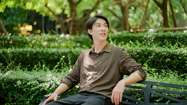 A young Asian man spending his free time getting some fresh air and relaxing on a bench in a public park.