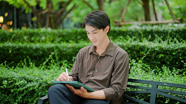 A young Asian man getting his work done on a digital tablet while spending time out of office and in a public park.