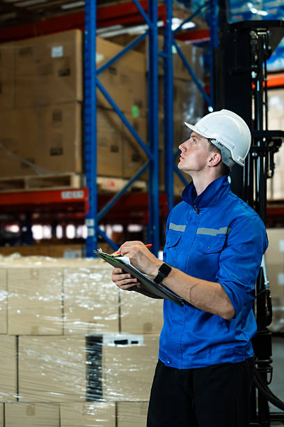 istock Male warehouse supervisor in blue uniform and white helmet holding clipboard observes ceiling area during warehouse inspection. Background filled with shelves and packed boxes. 2213998232
