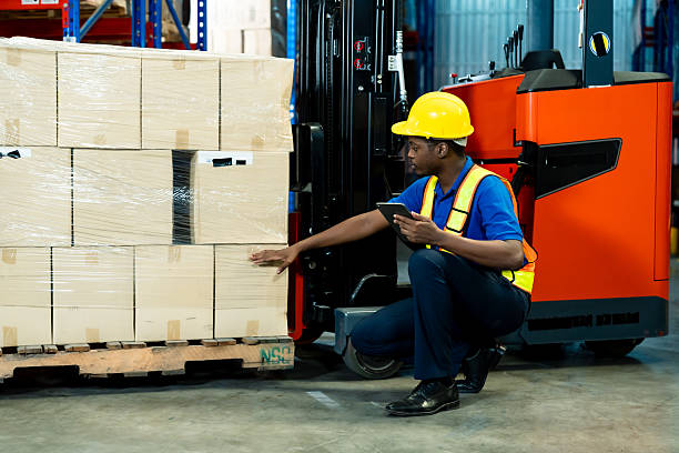 istock Warehouse worker kneeling beside stacked cardboard boxes on wooden pallet, inspecting packaging condition with hand, wearing yellow helmet and safety vest, holding tablet, forklift and shelves 2213998252