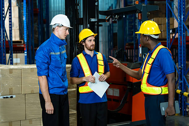 istock Group of warehouse workers wearing uniforms, safety helmets, and reflective vests logistics task, holding documents and tablet, standing near forklift truck inside distribution center full of boxes. 2213998259