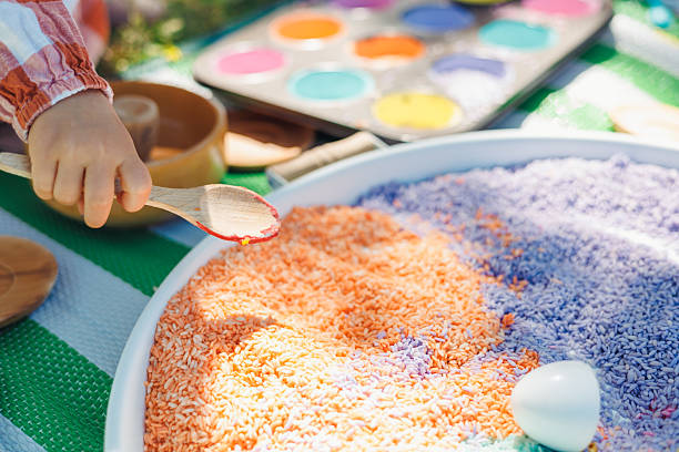 A hand reaches into a large tray filled with brightly colored rice during a sensory play session. This engaging activity takes place outdoors, fostering creativity and exploration among children.