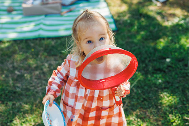 A young child in a checkered orange and white dress holds a red strainer, playfully shouting into it. The setting is a lively picnic scene in a park, with green grass and blankets spread out nearby.