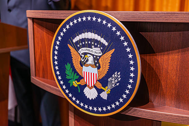 istock Image depicts a press conference at The White House, with focus on a blue lectern An eagle emblem, arrows and olive branch, stars, and stripes are present Indistinct figures of people likely in att 2214009927