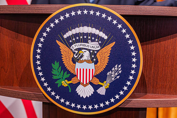istock Podium with US government emblem, featuring an eagle, E Pluribus Unum motto, shield, star stripes pattern, bundled arrows, olive branch, clouds, soft shadow, official style, no people 2214009929