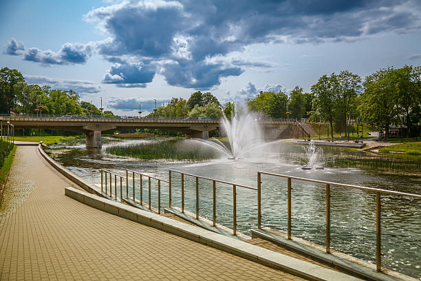 Riverside scene in Anyksciai, Lithuania, showcasing a bridge over a tranquil river with fountains. A paved walkway runs alongside, inviting leisurely strolls beneath picturesque clouds