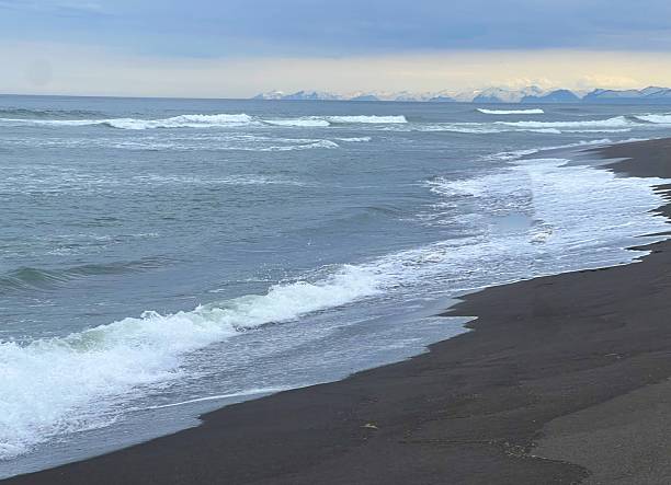 istock Serene ocean waves lapping dark sandy shore under cloudy sky with distant mountains 2214016257