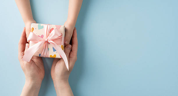 istock Close-up of hands exchanging a gift wrapped with a pink ribbon on a light-colored surface, conveying themes of giving, love, celebration, and appreciation 2214025567