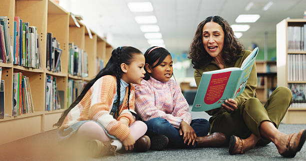 Girl, teacher and book with reading at library for development, learning or study with guide. School kids, storytelling and woman with help on floor, education and language for knowledge at academy