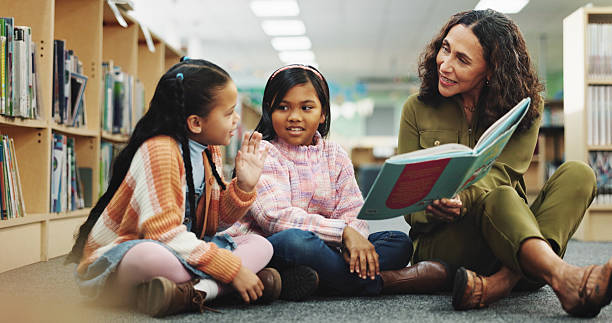 istock Girl, teacher and teaching with reading book at library for development, learning or study with guide. School kids, storytelling and woman with help, education and language for knowledge at academy 2214027170