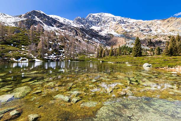 It is springtime in the mountains surrounding the sanctuary of Oropa, a UNESCO World Heritage Site. On this beautiful day, the sun is shining and nature is awakening from its winter hibernation. Here is a beautiful small mountain lake, reflecting the surrounding mountains and woods, still covered with fresh snow.