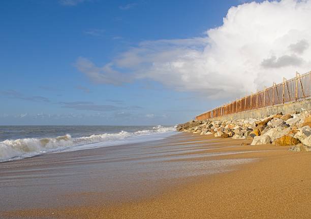 Ocean waves rolling onto a sandy beach with a rusty fence and rocks in the background under a cloudy sky