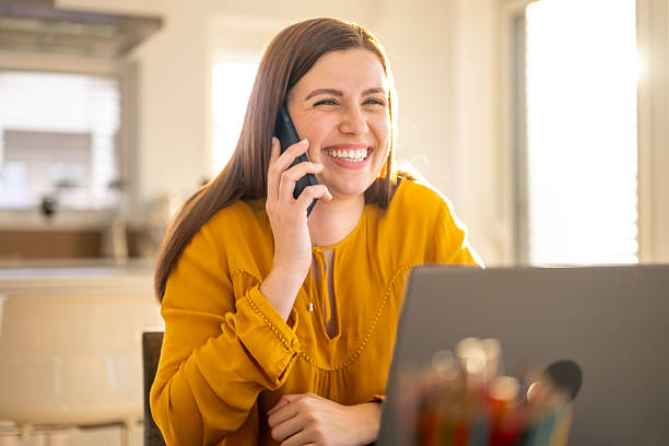 istock Smiling Young Woman in Bright Kitchen Talking on Phone While Working on a Laptop 2214031293