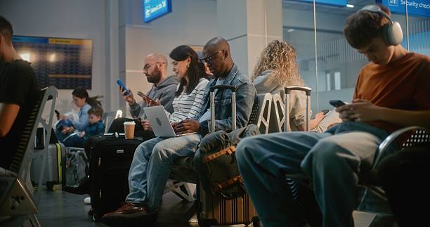 Crowded International Airport Terminal: Diverse People, Passengers Sitting in Departure Lounge of Airline Hub, Waiting for Airplane Flight, Using Phones. African American Man Working Online on Laptop.