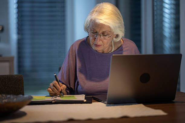 istock Senior Woman Taking Notes While Working On Laptop In Evening Home Office Setting 2214033830