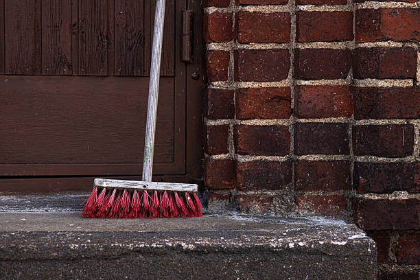 Broom with red bristles leaning against rustic brick wall outdoors. Red bristled cleaning mop stands upright next to a textured brick wall and wooden door