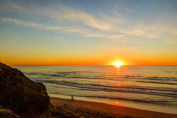 A vibrant sunset over the ocean, with the sun dipping below the horizon. Silhouettes of two people walk on the beach near a cliff.