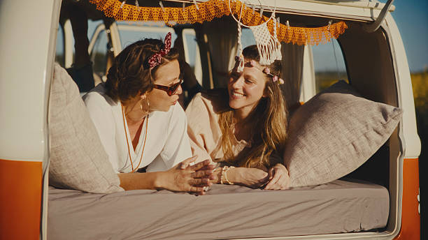 Two Smiling Women Lounging on a Cozy Bed Inside a Vintage Van,Sharing Laughter and Enjoying a Relaxing Moment During their Adventurous Travel or a Vibrant Festival