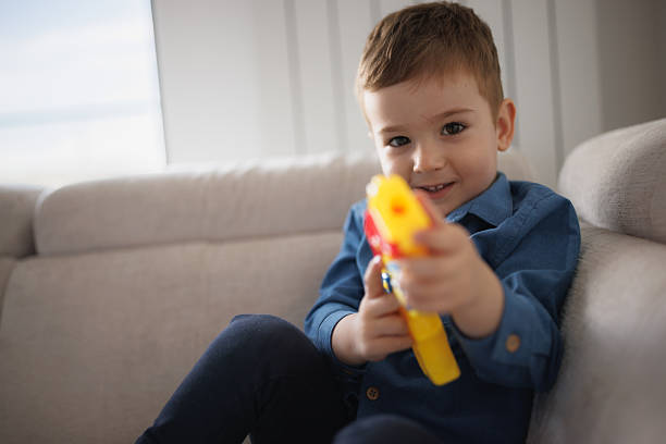 Cute little boy pretending to be a hero with a plastic toy gun while lounging on the living room sofa.