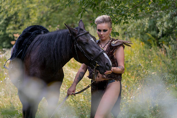A woman dressed in warrior-like clothing gently interacts with a black horse. They are surrounded by lush greenery, capturing a serene moment in nature.