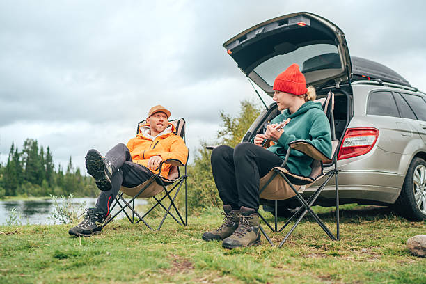 istock Peaceful Norwegian camping scene with father and son by lakeside. They relax in chairs while son plays ukulele, surrounded by nature, laughing and enjoying moment together near their car in wilderness 2214068784