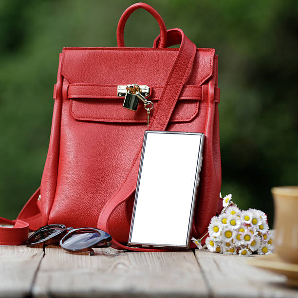 Red leather handbag with a metal lock detail sits on a wooden table. Nearby, a smartphone with a blank screen leans against the bag. A small bouquet of daisies (Bellis perennis) adds a touch of nature, accompanied by a pair of sunglasses. A cup and saucer are partially visible, suggesting a casual, outdoor setting. The background features a blurred, green garden environment.
