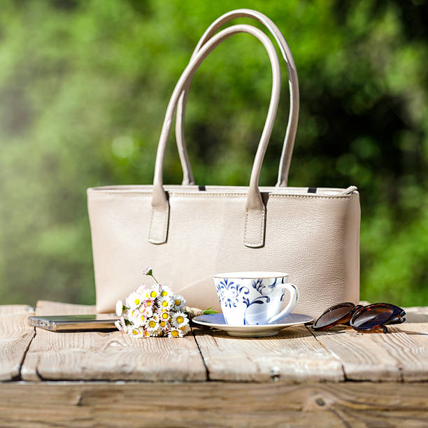 Beige handbag sits on a rustic wooden table, accompanied by a small bouquet of daisies (Bellis perennis), a smartphone, sunglasses, and a decorative cup with a matching saucer. The cup features a blue floral design. The background shows a blurred garden, suggesting an outdoor setting. Mother's day or Women's day or greeting card background with copy space.