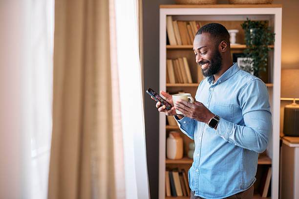 istock Smiling man drinking coffee and using smartphone near window at home 2214076798